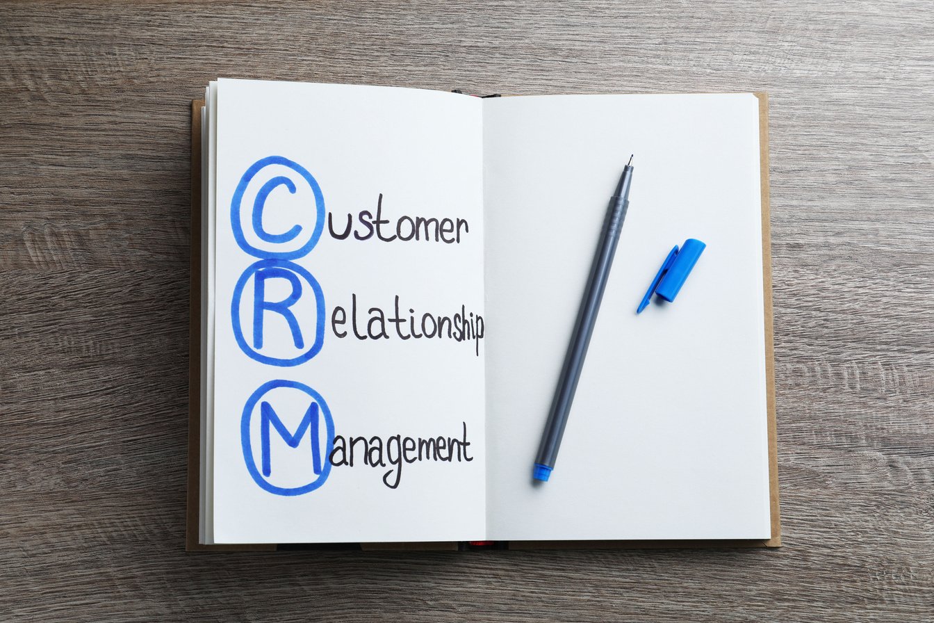 Notebook with Abbreviation CRM (Customer Relationship Management) and Pen on Wooden Background, Top View